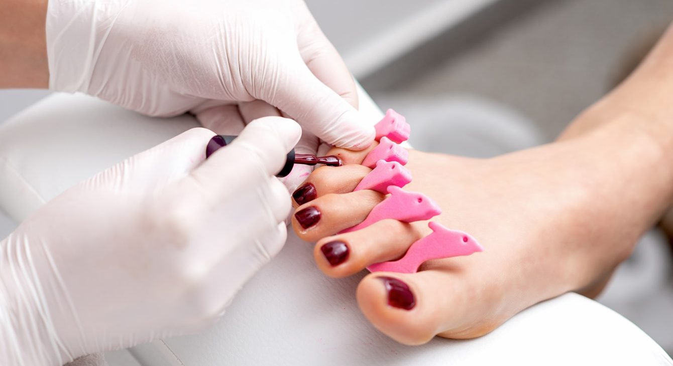 manicure-master-is-painting-female-toenails-with-maroon-nail-polish-by-brush-wearing-white-gloves